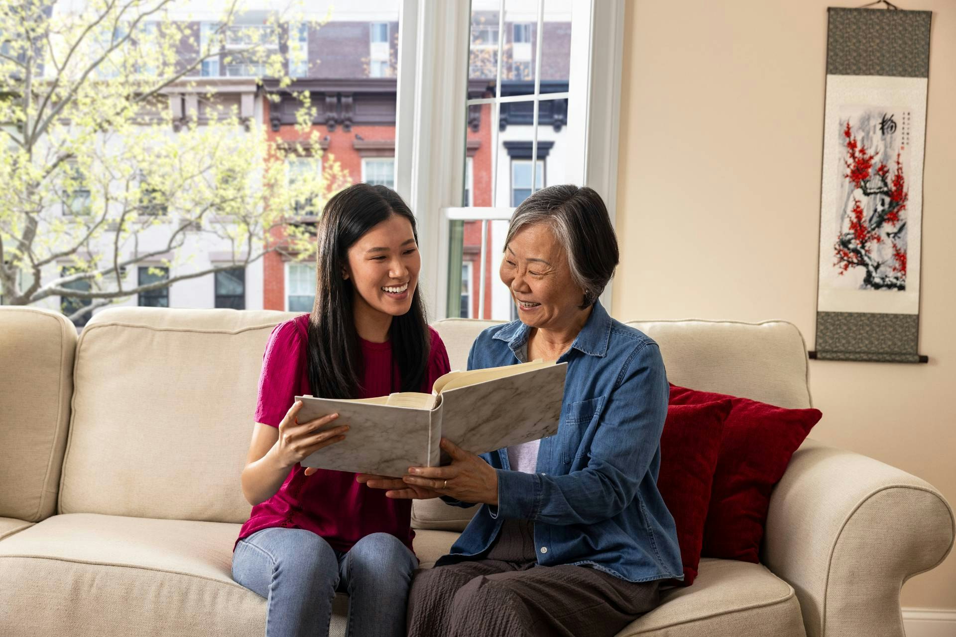 A woman and her adult daughter sit on the couch looking at a book together