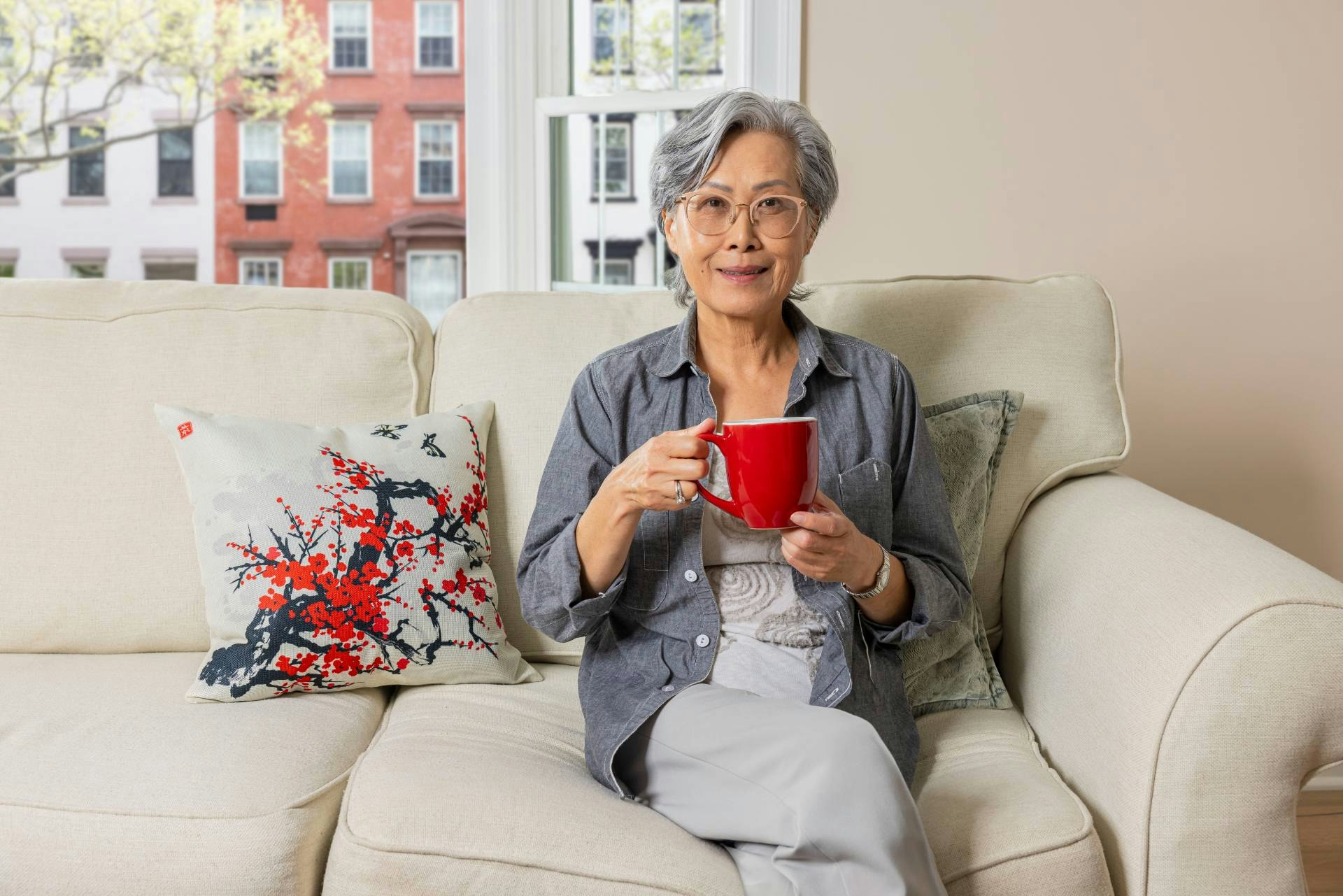A woman sits on a couch. She wears glasses and holds a red mug