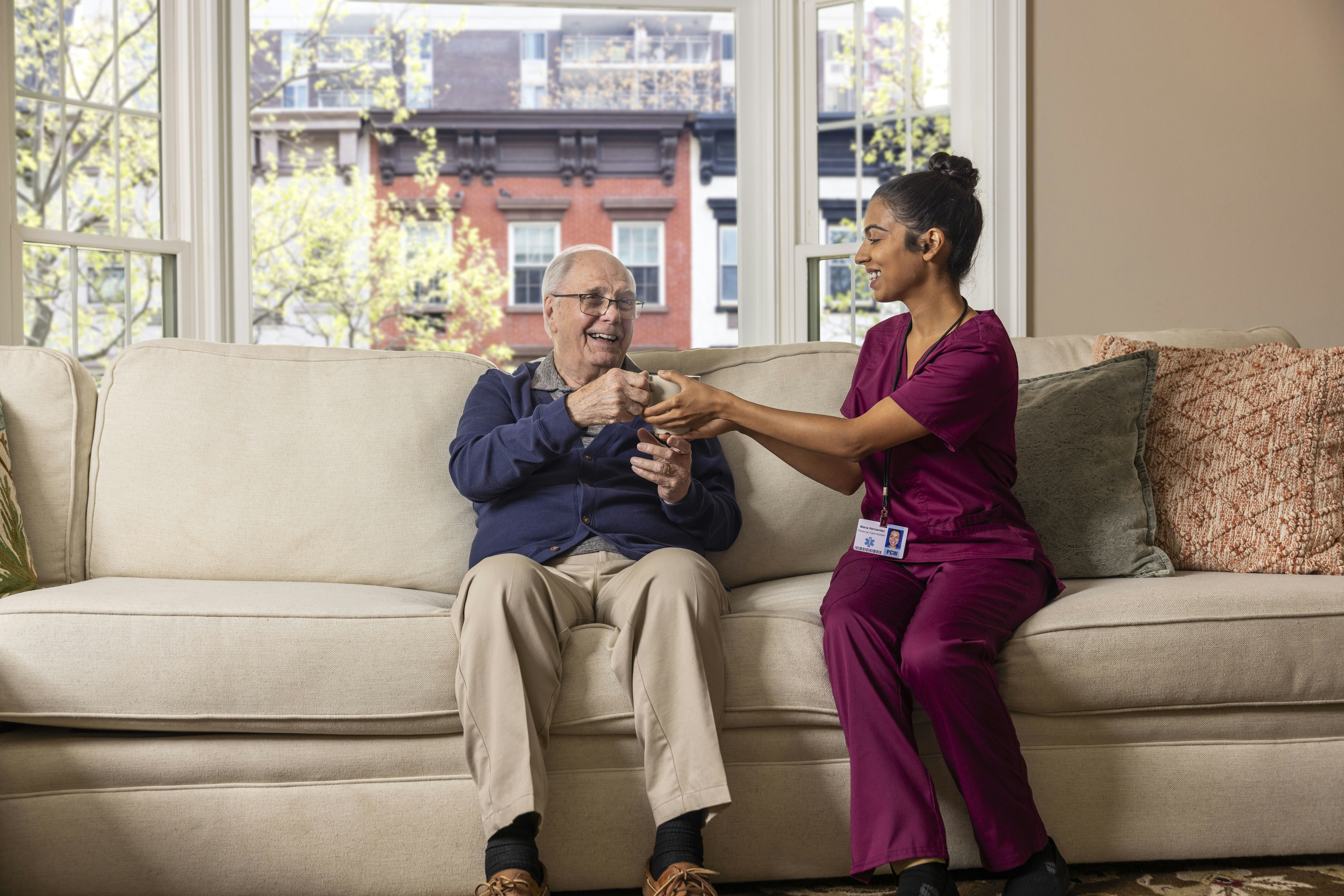 A nurse in magenta scrubs sits on a couch with an older patient and hands him a cup of tea