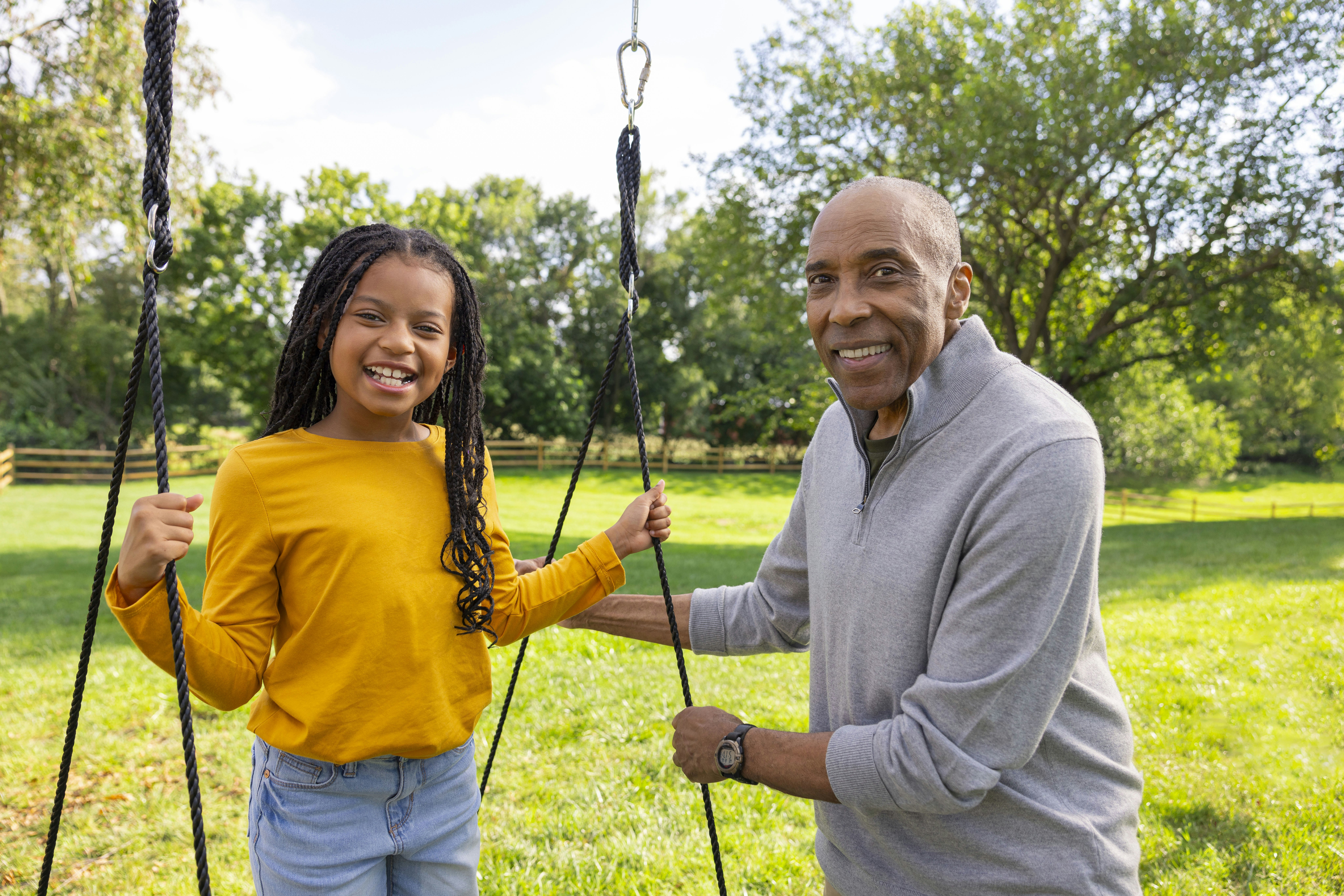 A grandfather and granddaughter smile at the camera while he pushes her on a swing