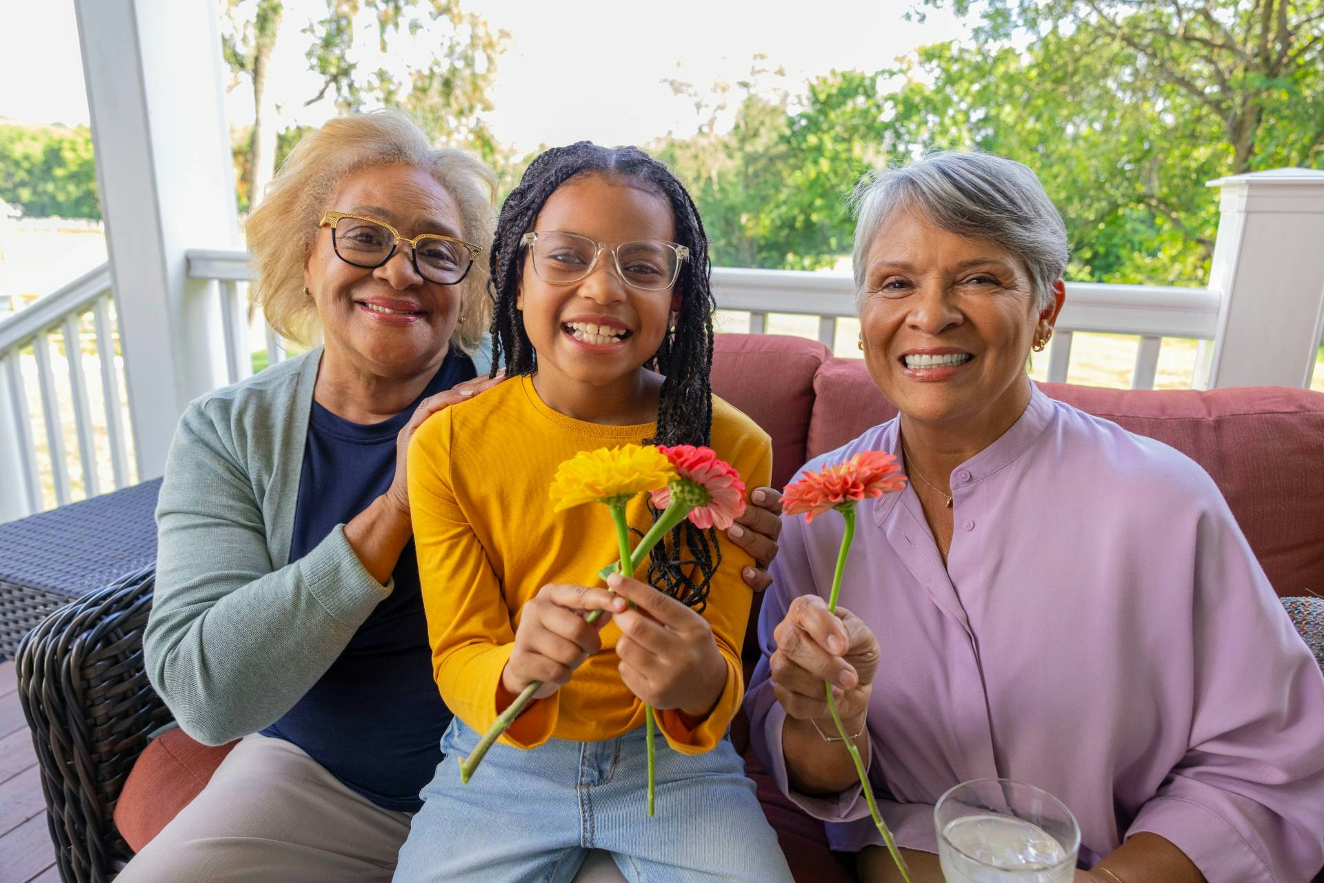 Two older women and a young girl sit on a bench on a porch. They smile at the camera