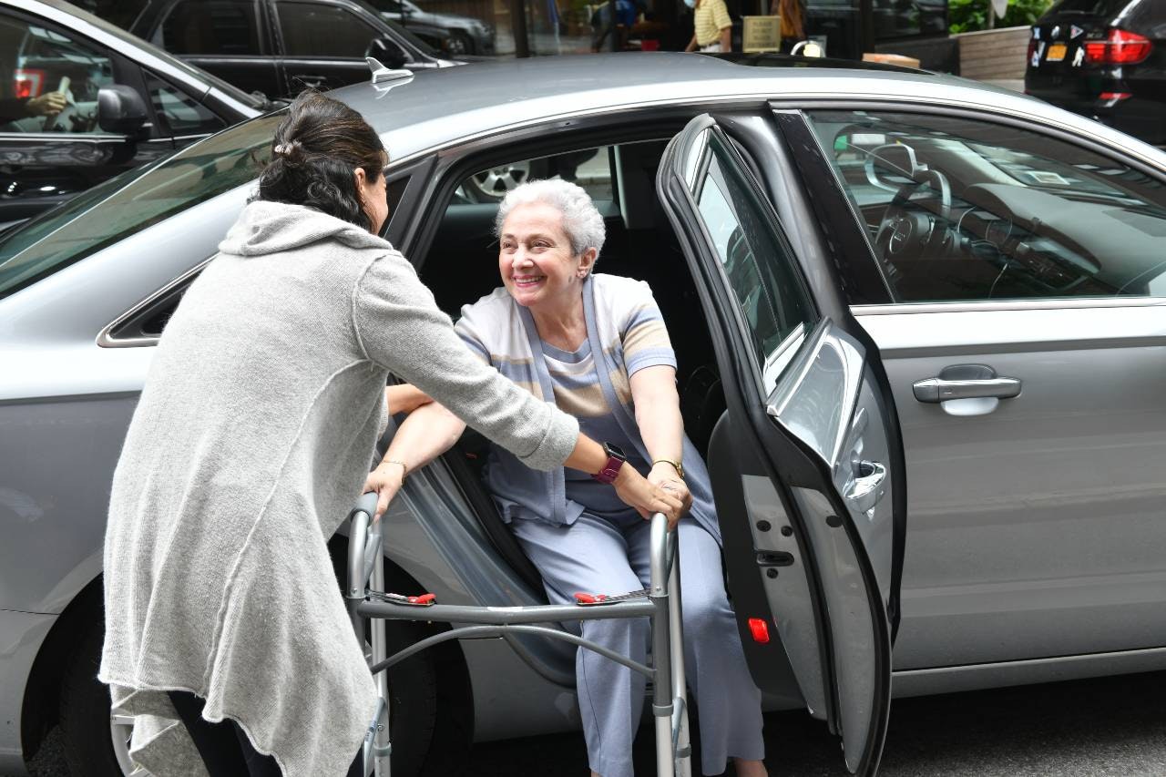 A woman with short gray hair is helped out of a car, using a walker.