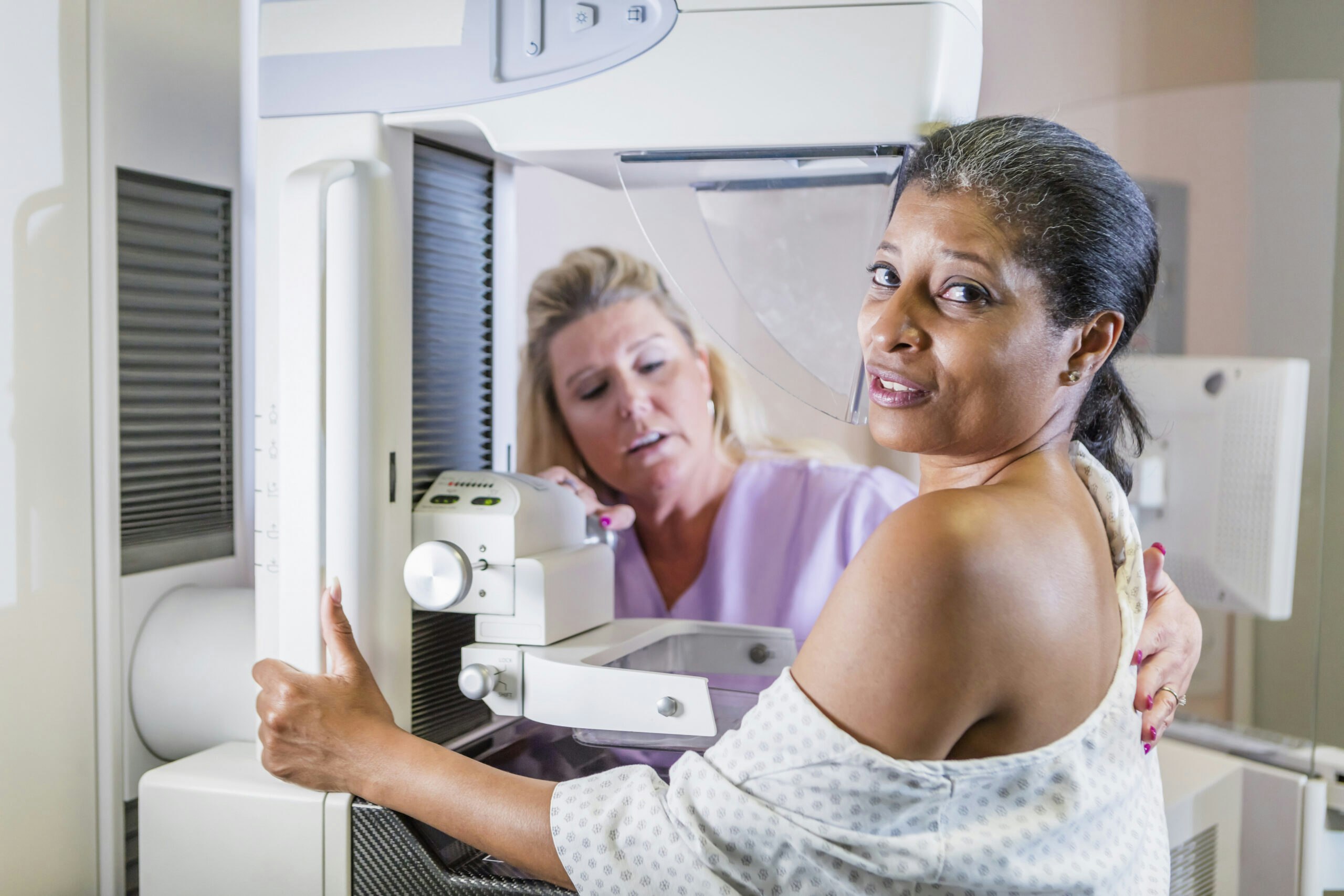 A mature African-American woman in her 40s wearing a hospital gown, getting her annual mammogram. She is being helped by a technologist, a blond woman wearing scrubs. The focus is on the patient, who is looking toward the camera.