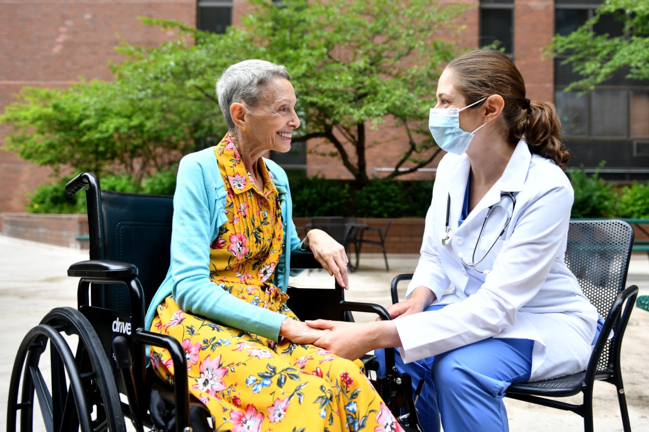 A resident in a wheelchair speaks with a masked nurse in a courtyard.