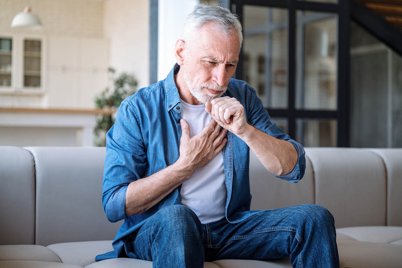 Mature man suffering from dry cough and chest pain while sitting alone on sofa in living room.