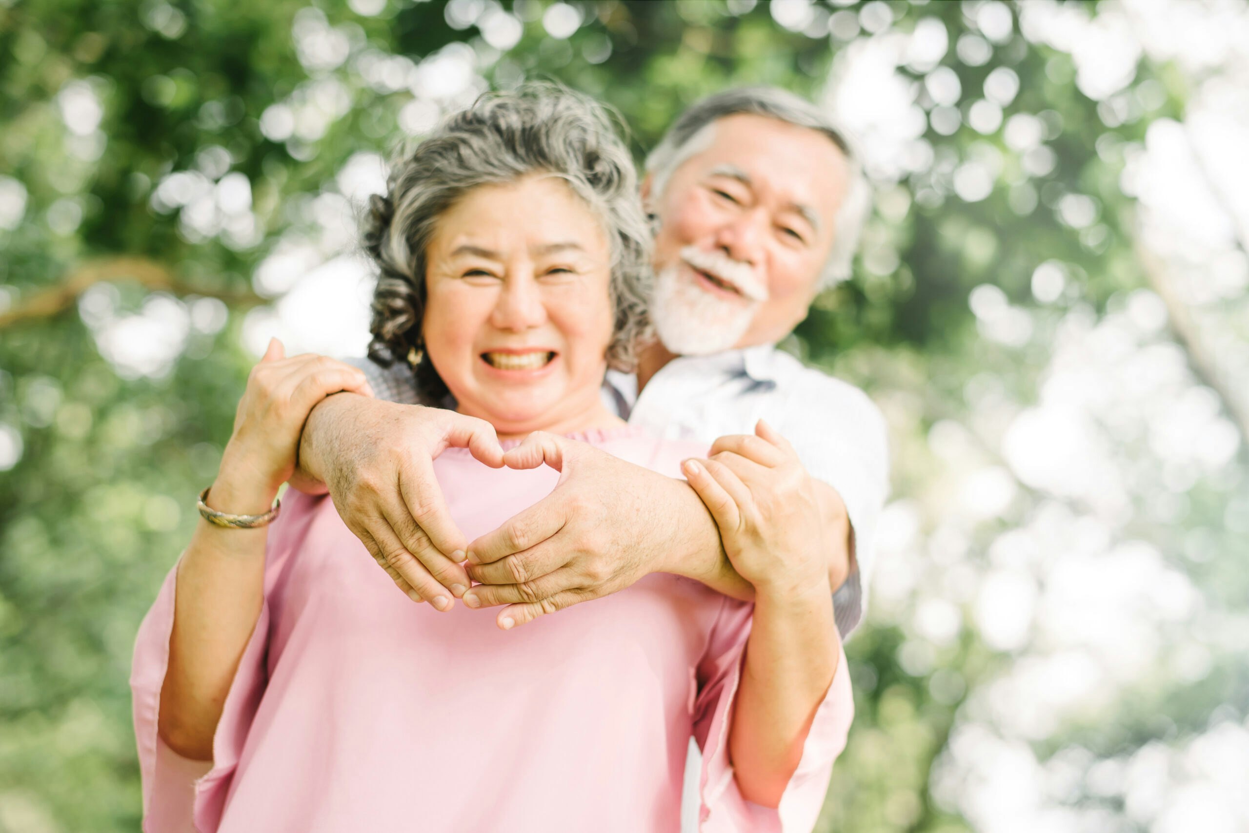 An older couple smiles at the camera. The man stands behind the woman, arms over her shoulders, and makes a heart symbol with his hands.
